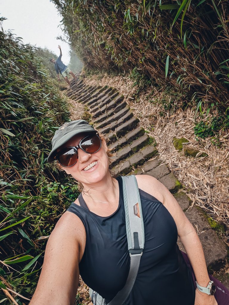 Sooz taking a selfie while walking down stairs amongst greenery, with Ganesh waving from further up the path behind.