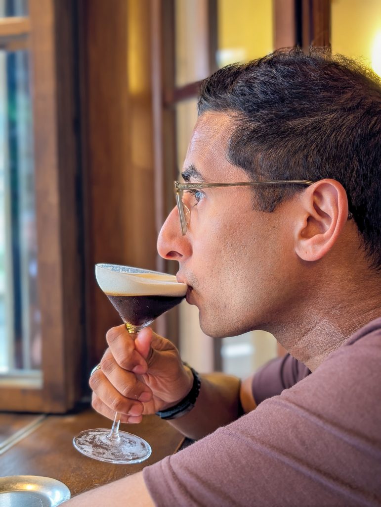 Ganesh sipping a brown and frothy drink in a long cocktail glass and looking very refined.
