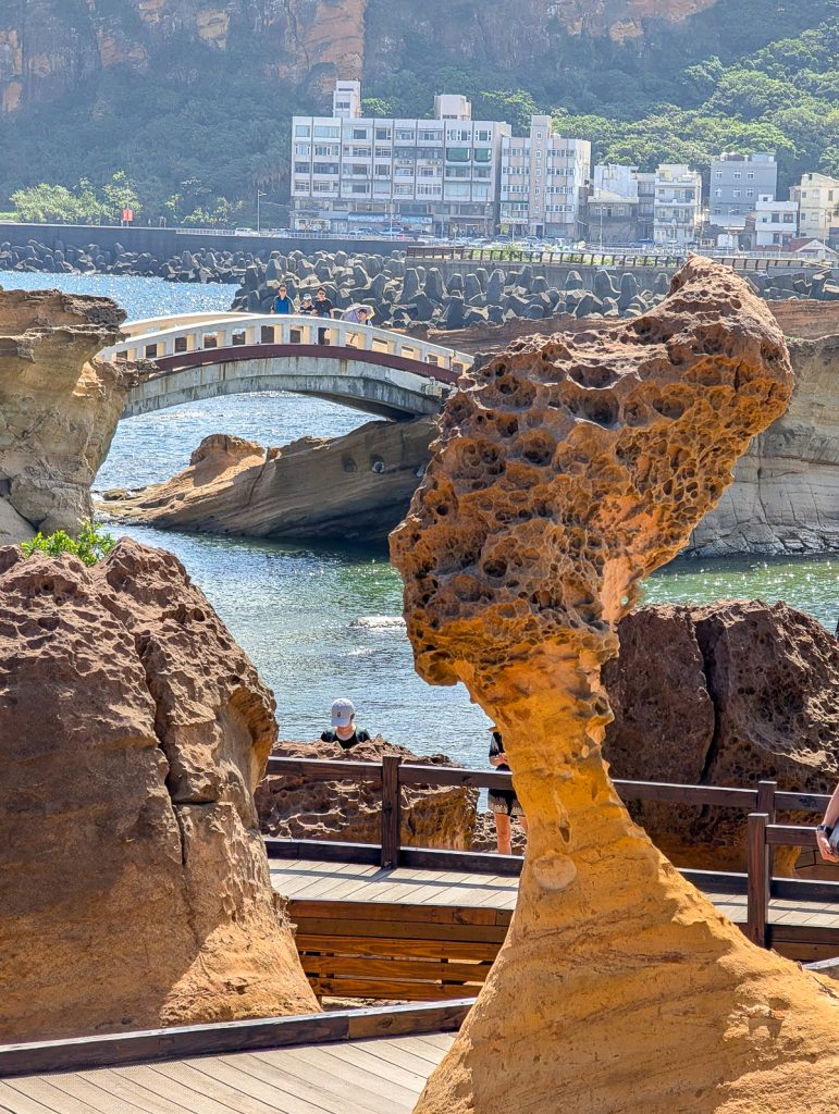 A sandstone rock formation that looks like a head on a neck, with more rocks, a bridge and some people in the background.