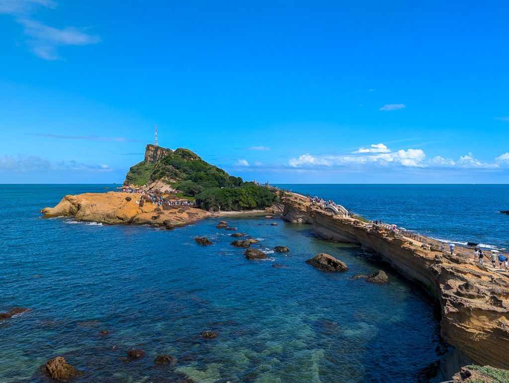 A rocky isthmus leading out to a green-topped sandstone headland with a white and red lighthouse on the tip. The sky and sea are beautifully clear and blue.