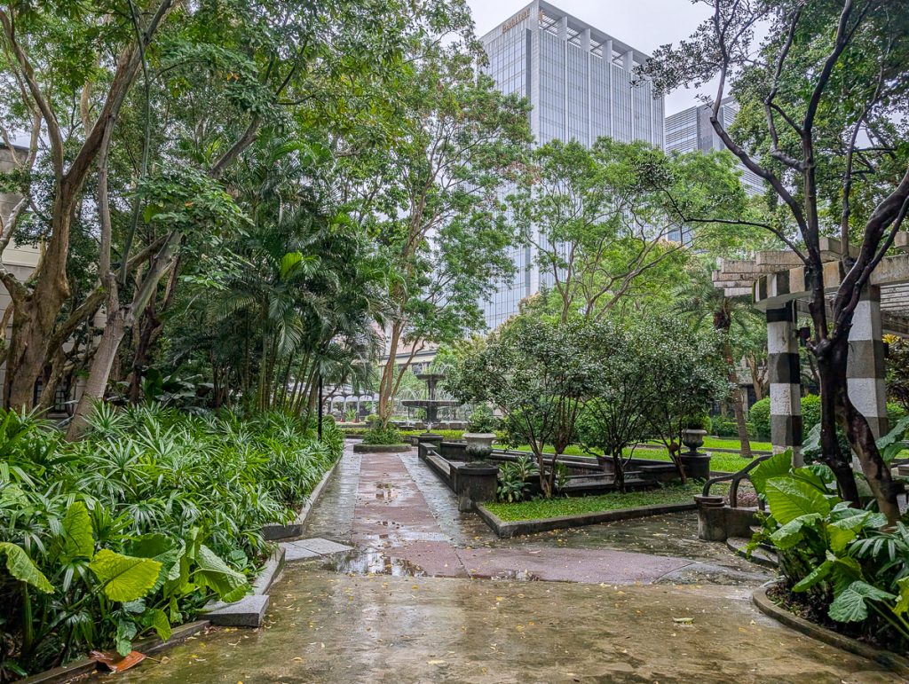 A lush green park with pathways and a fountain, with city buildings peeking over the top.