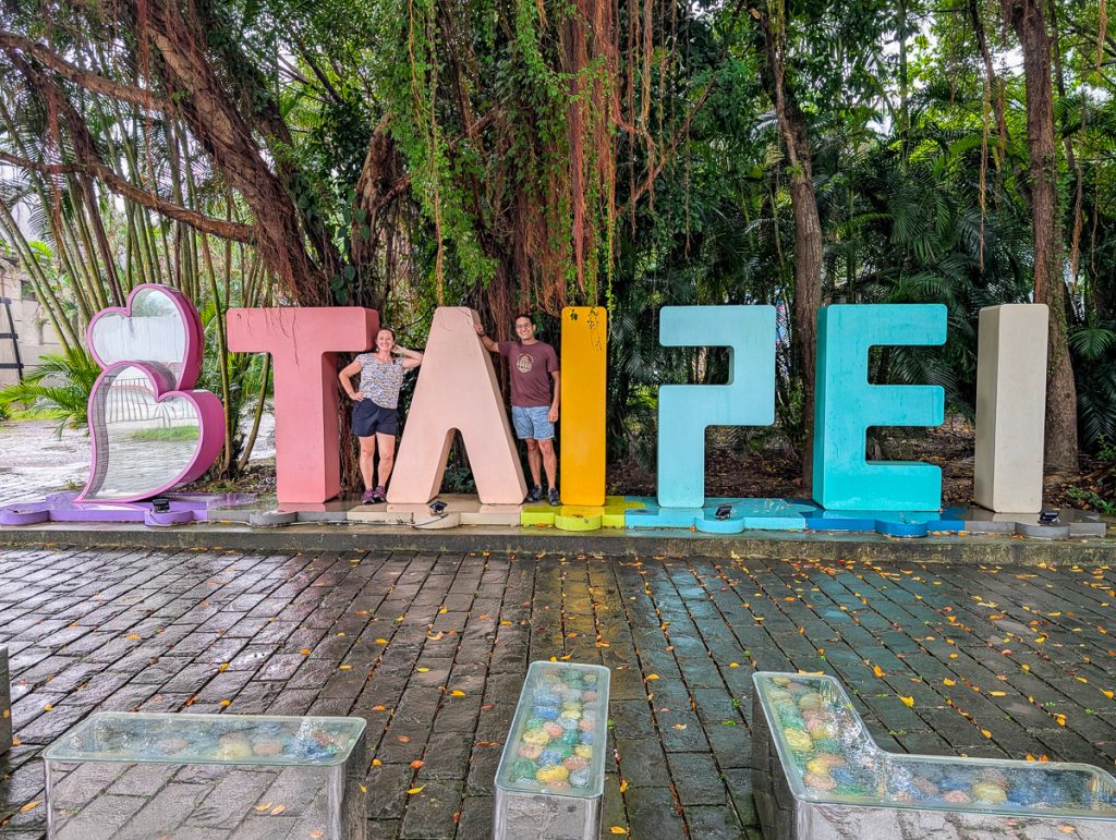 A man and woman posing with giant colorful letters spelling out Taipei with trees behind.