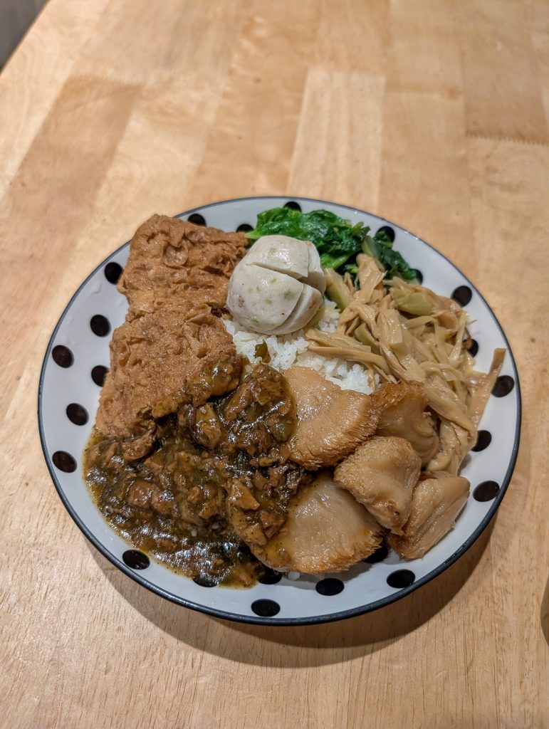 A white plate with black spots heaped up with white rice, greens and a variety of brown foods.