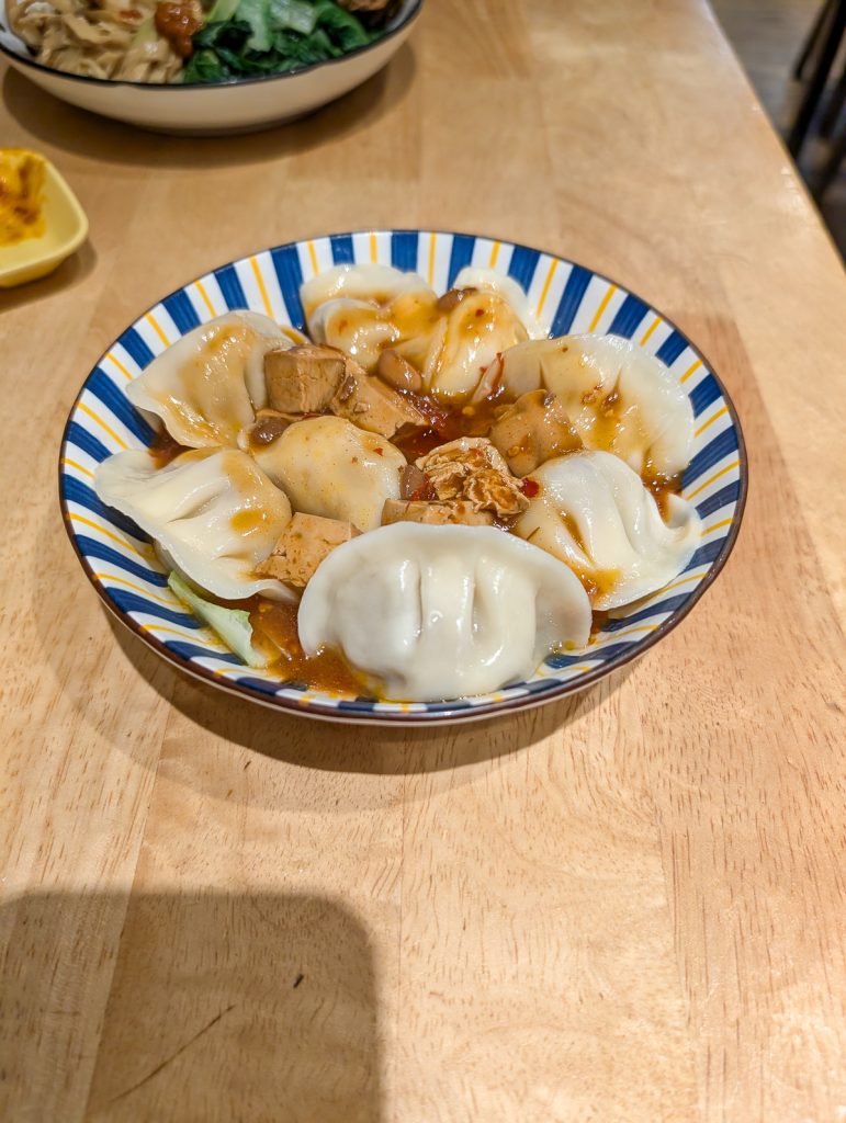 A striped white, yellow and blue bowl filled with dumplings, tofu and a sticky-looking sauce.