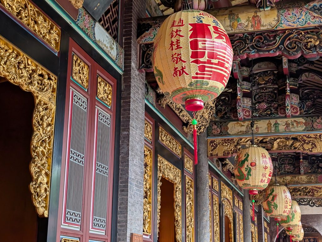 A close up of paper lanterns with writing and images hanging from an ornate temple ceiling with gold and paintings all around.