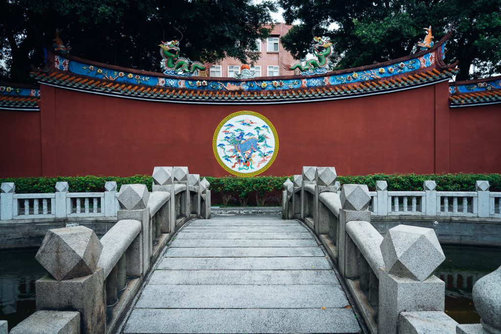 A bridge with geometric stone railings leading to a red wall with a circular painting of a dragon on it, and more decorative dragon statues on top.