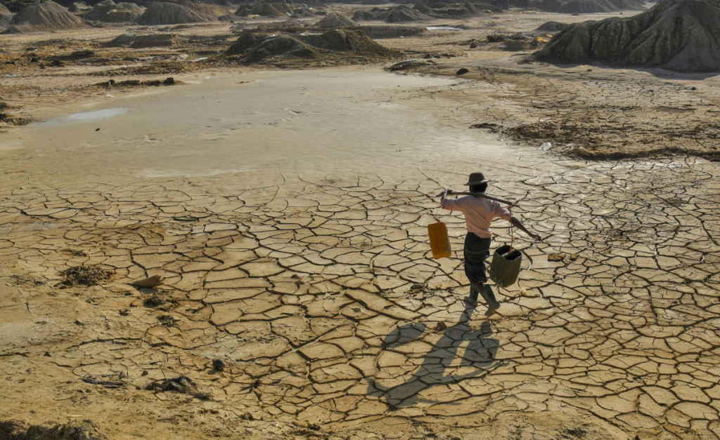 A man walking across dry mud flats while balancing a pole holding two buckets on his shoulders.