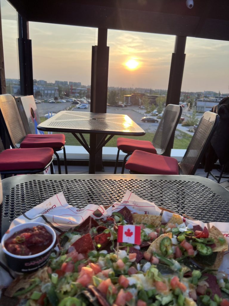 A close-up shot of a plate of nachos with a Canadian flag in them, and a view across tables to a window and a low setting sun behind.
