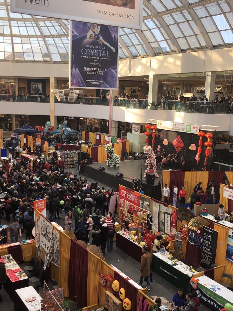 A crowded mall with small stalls and a stage with people performing to a crowd as seen from a second-floor indoor balcony.