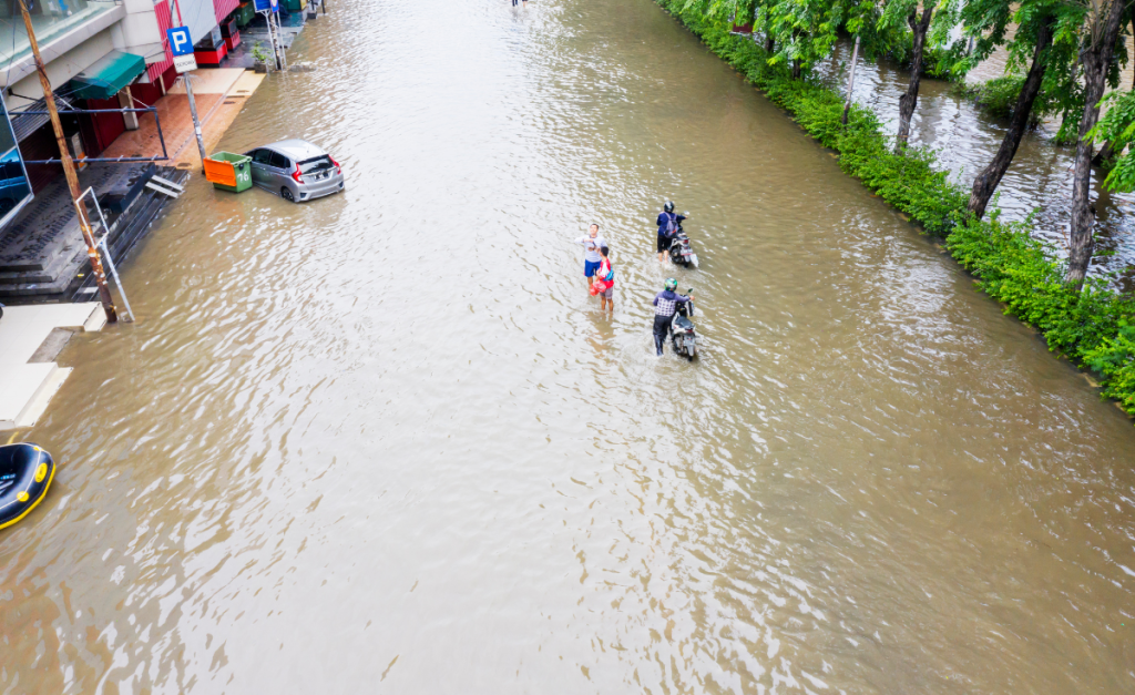An aerial photo of a flooded road with some people standing in the water or pushing mopeds in front of buildings.