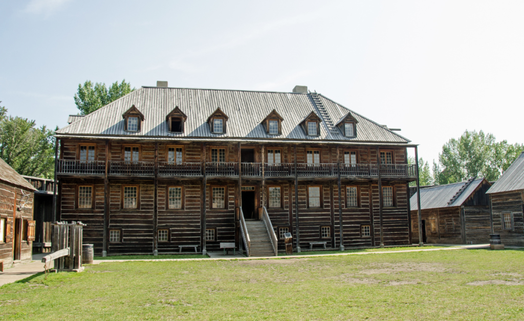 A three storey wooden building on green grass, with smaller buildings around it.