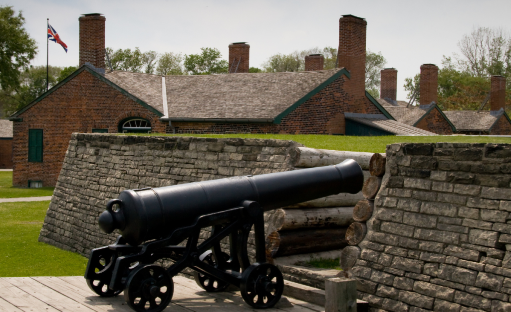 A cannon on wheels next to a stone wall with a red brick building in the background flying a Union Jack flag.