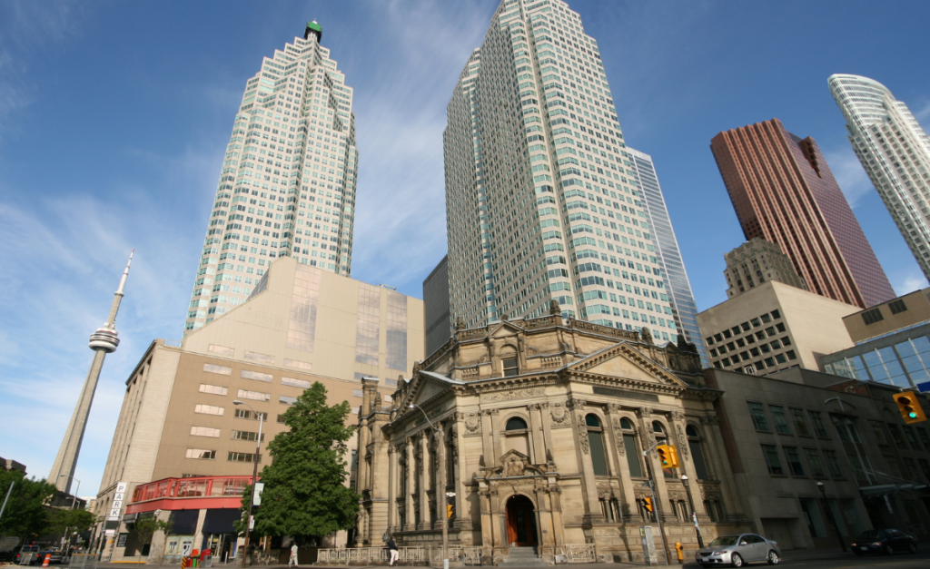 A city street with a sandstone building in the foreground and tall skyscrapers and a tower in the background.