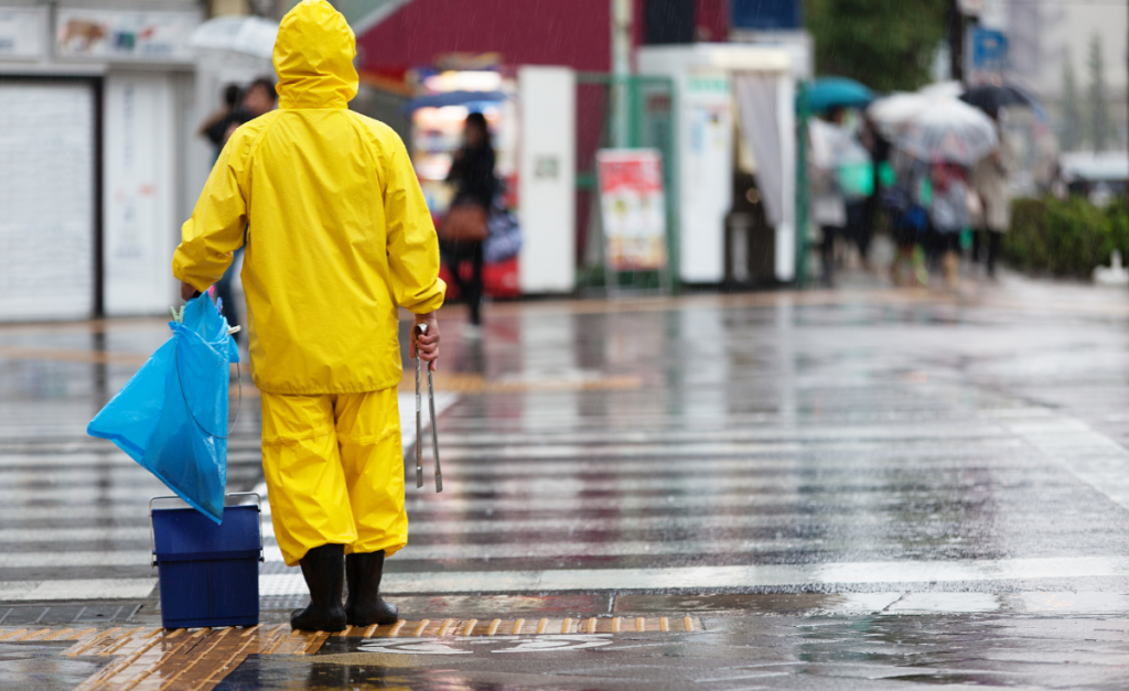 A person in a complete yellow waterproof outfit holding a blue plastic bag in rain next to a road crossing.