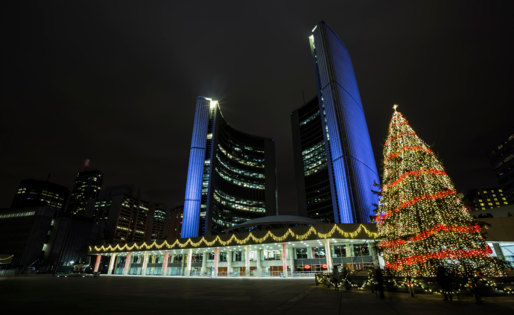 Two curved tall buildings lit up with blue light, above a smaller building with sparkly lights and a big Christmas tree lit-up at night.