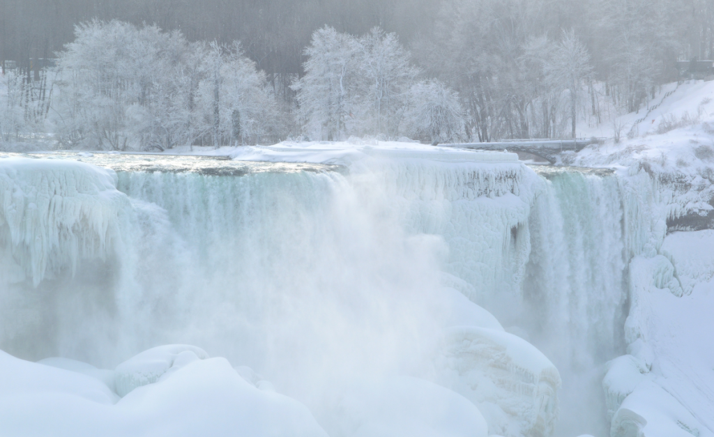 A large waterfall almost completely frozen over with snow on the banks and trees in the background.