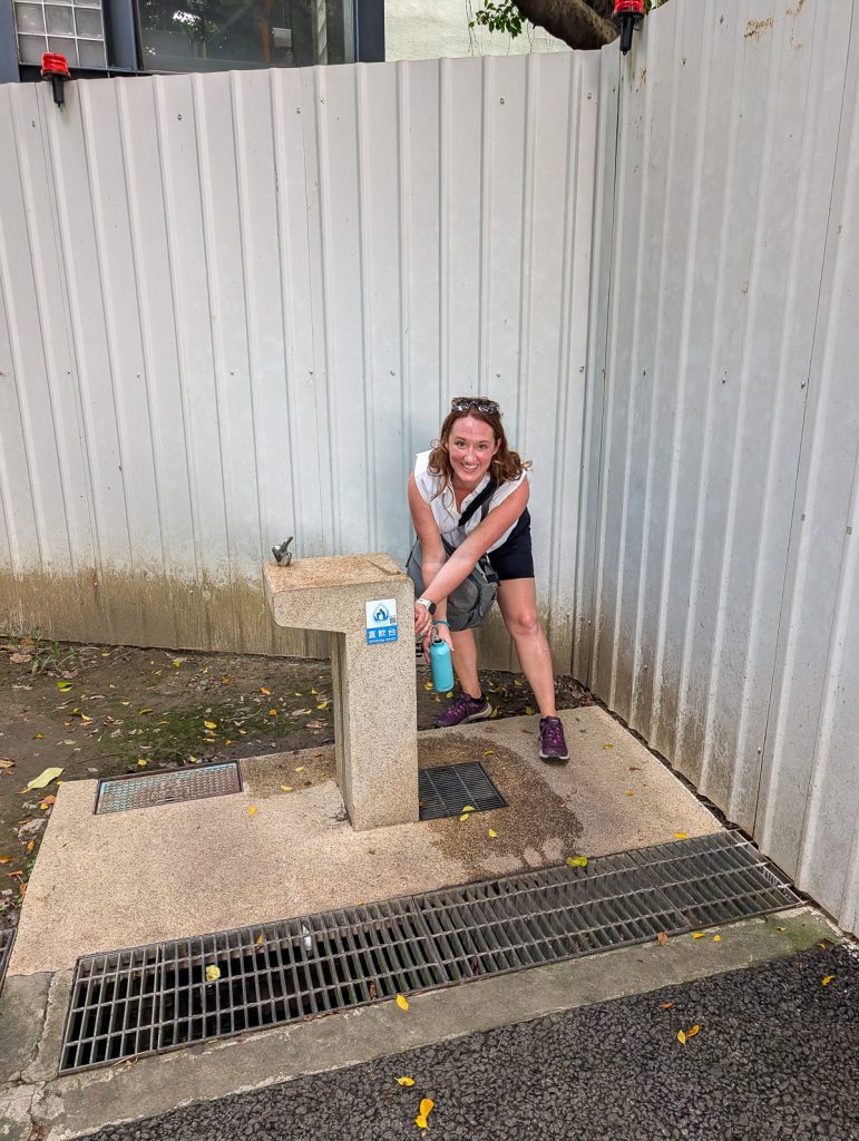Sooz filling up a blue water bottle at a water refill station with a white fence behind her.