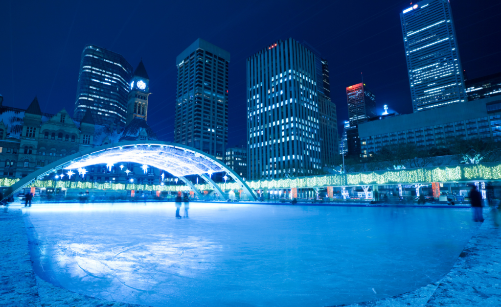 An ice-skating rink at night with an arched roof at one end, lights around the edges and skyscrapers in the background.