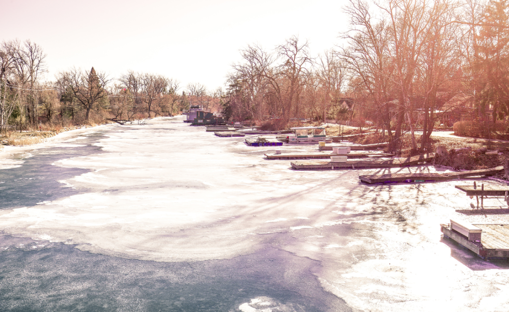 An almost frozen over river with docks jutting out from the bank on a hazy day.