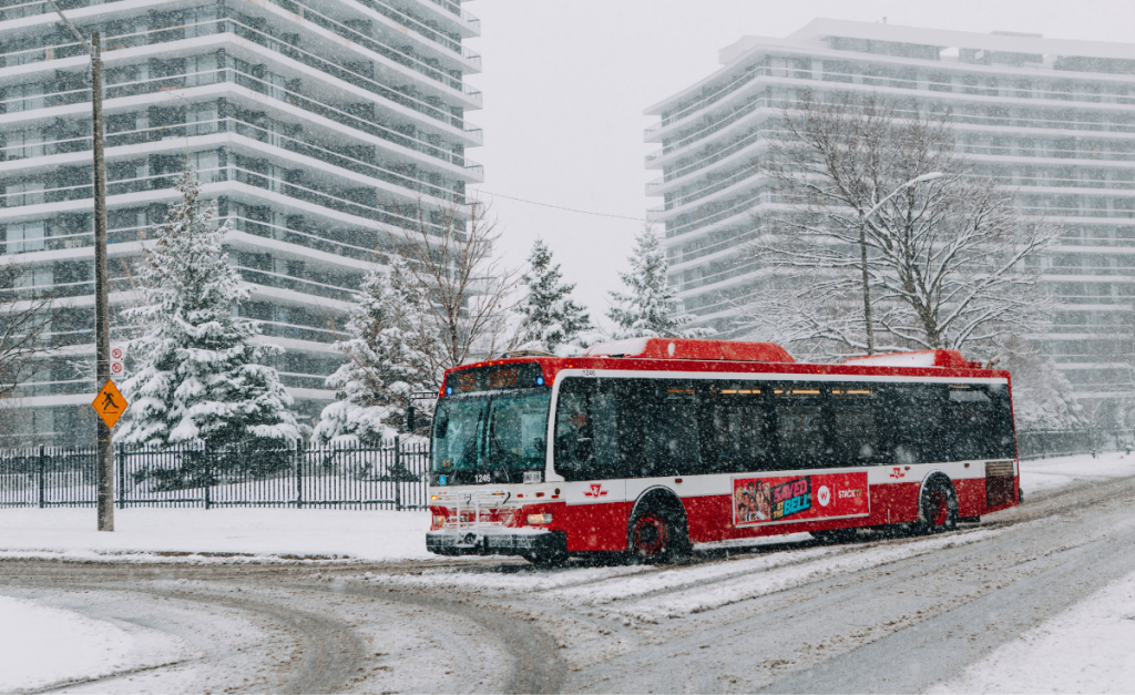 A red and white bus driving on a snowy road with apartments in the background.