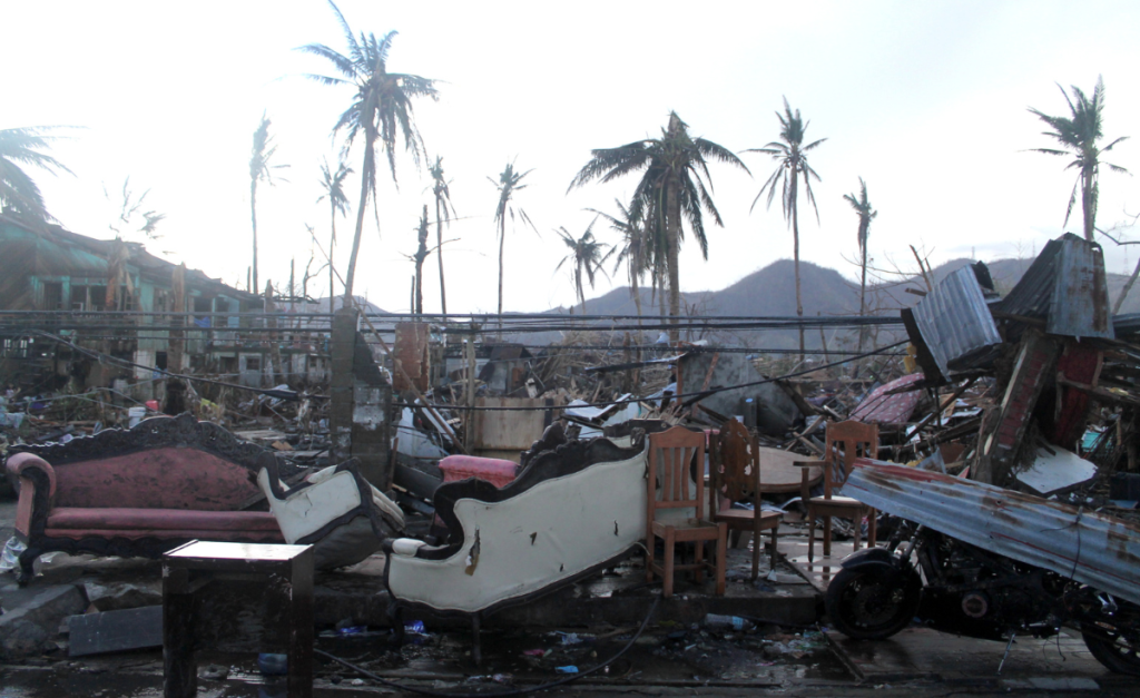Piles of furniture and broken pieces of metal with battered looking palm trees and buildings in the background.