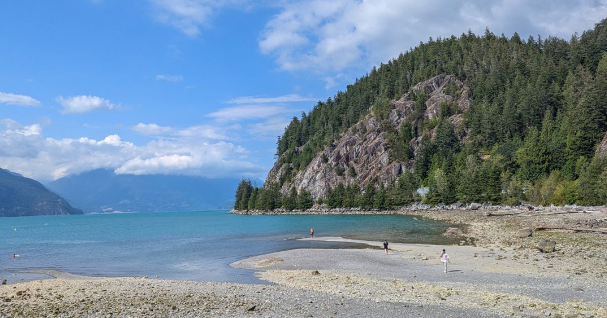 A landscape photo of a rocky beach with a few people on the shore and in the water, with forested mountains in the background and a blue sky with a few white clouds above.
