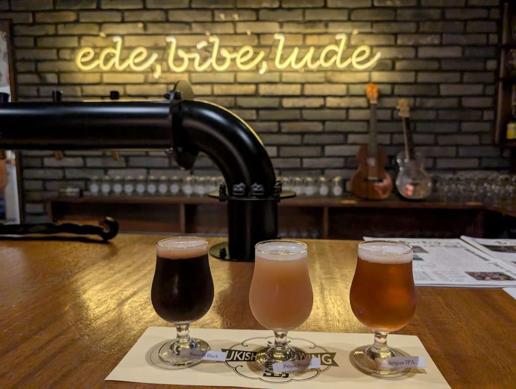 A close up of three glasses of beer with labels sitting on a bar with a pipe, brick wall and neon lights behind.