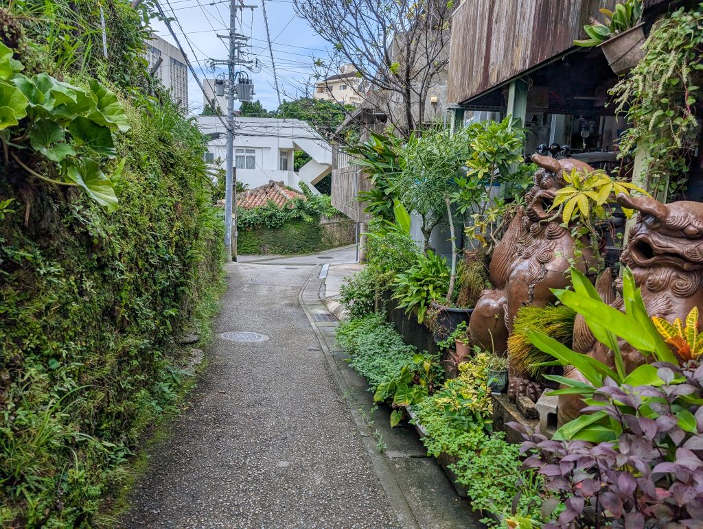 A small alleyway filled with greenery and two big brown sculptures, leading to a street with buildings. 