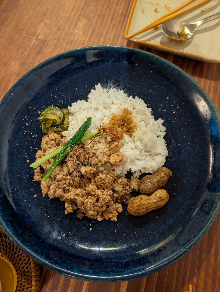 An overhead close up shot of a shiny blue bowl with white rice and brown food with some whole peanuts on the side.