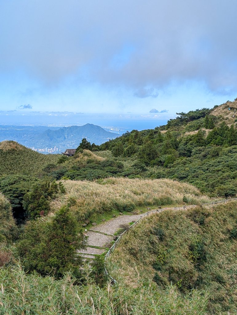 A walking path on a green hill with mountains and buildings far away in the distance below.