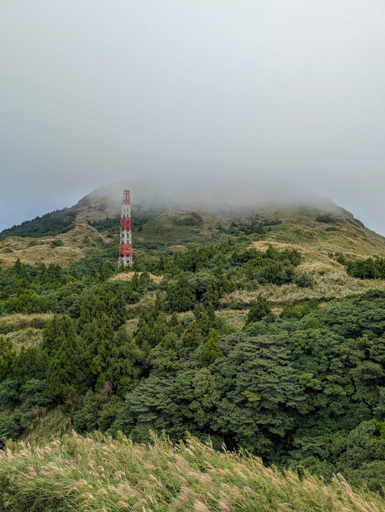 A small red and white tower on the side of a green hill with fog covering the peak.