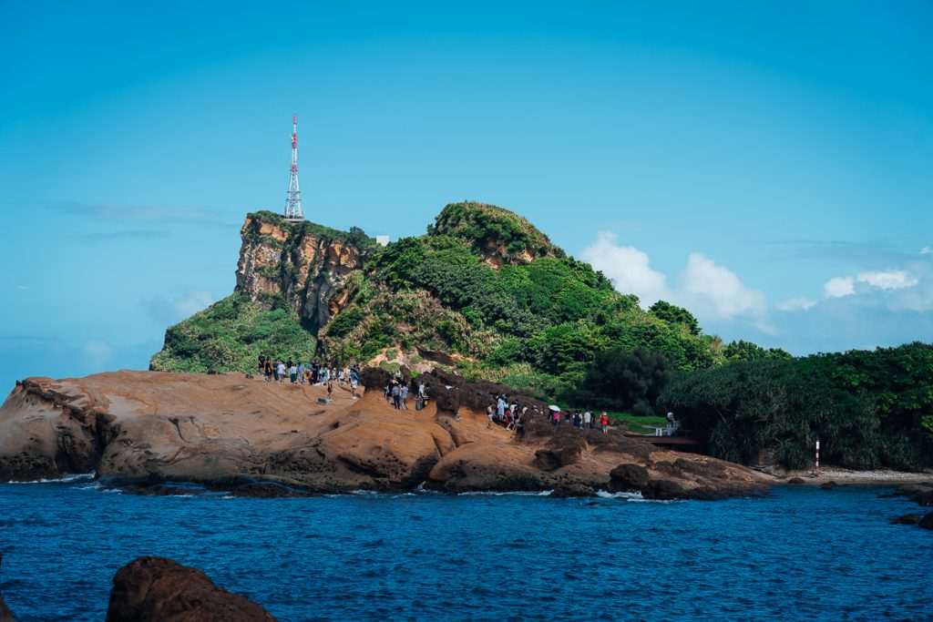 A rocky headland with lots of people on the rocks at the foreground and greenery and a lighthouse in the background.