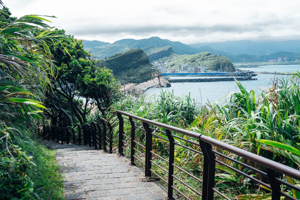 Paved steps with a fence next to greenery on a headland with hills, buildings and clouds in the background.