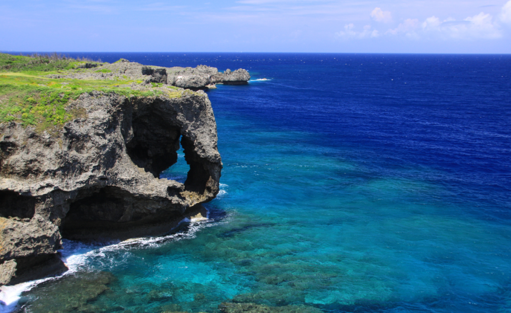 Rocky cliffs with grey stone formations topped with grass next to a  deep blue sea under blue skies.