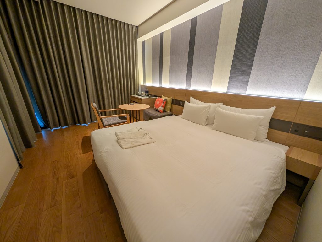A big white bed in a minimalistic hotel room with the curtains closed.