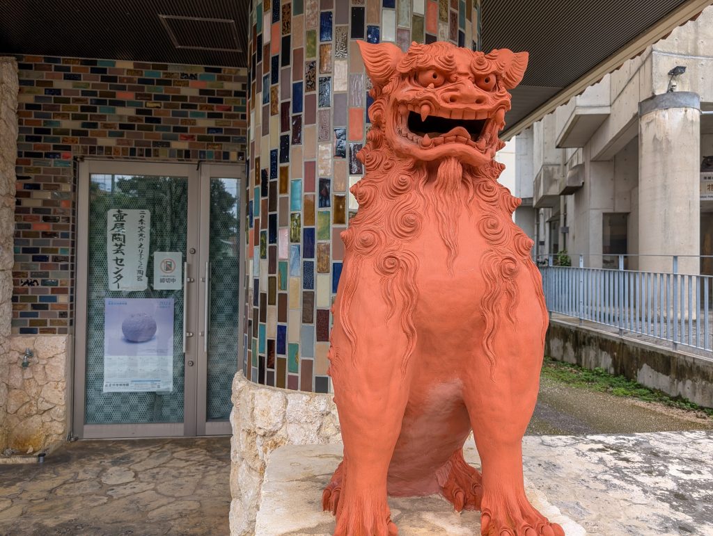 A large orange statue of a lion dog sitting in front of a building with colorful tiles on the exterior.