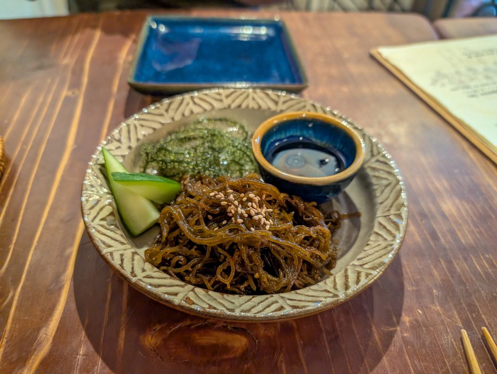 A close up shot of a dish holding brown noodles, cucumber slices, seaweed and soy sauce.