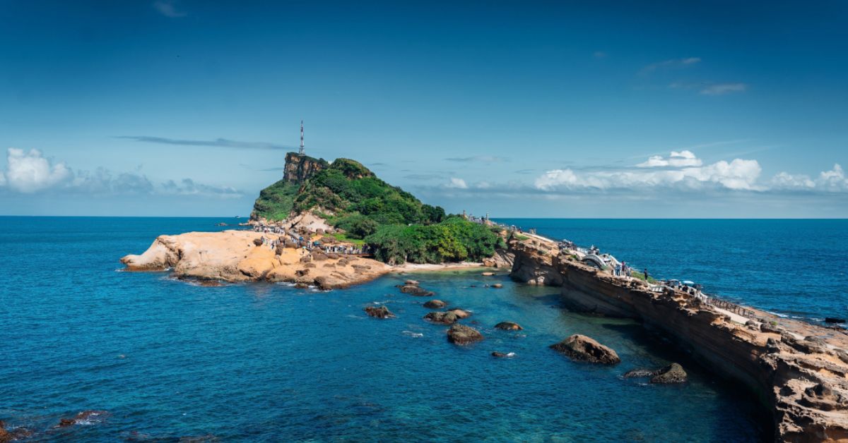 A landscape photo of a small rocky headland jutting into the ocean, with greenery and a small lighthouse at the end.